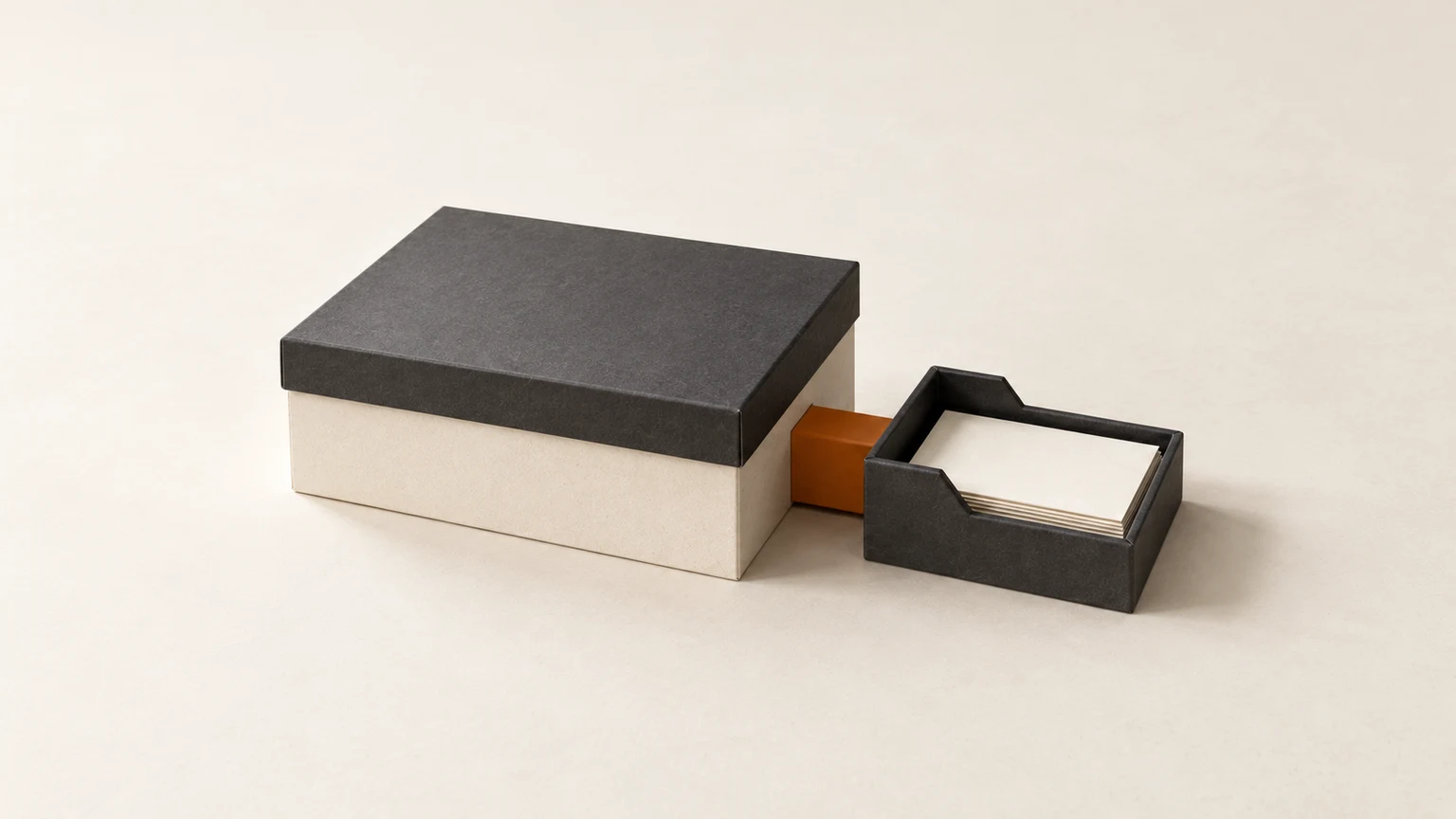 A closed cream ledger box with a graphite lid, joined by a short burnt-orange connector to a smaller graphite tray of blank cards.
